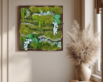 Peut inclure: Une œuvre d'art murale carrée composée d'un arrangement dense de mousse verte préservée aux textures et nuances variées. La composition comprend de petites plantes succulentes, de la verdure tombante et des accents floraux blancs délicats.