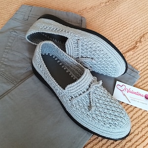 Puede incluir: Un par de mocasines de ganchillo gris claro con suelas negras, colocados sobre un pantalón gris. Los zapatos tienen un patrón texturizado y un detalle de borla. También se ve una pequeña tarjeta con el nombre "Valentina".