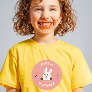 May include: A yellow t-shirt with a round graphic featuring a white rabbit peeking out of a hole, with the text "MADE IN AMSTERDAM". The t-shirt is worn by a person with curly hair and a messy face.