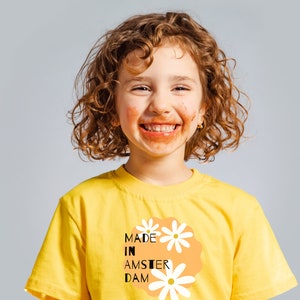 May include: A young person with curly hair smiles, wearing a yellow t-shirt. The shirt features the text "MADE IN AMSTERDAM" with daisy graphics on an orange background. The person's face is smeared with a brown substance, suggesting they are enjoying a treat.