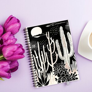 May include: A spiral-bound notebook with a black cover featuring a white and beige desert landscape design. The image includes a full moon, cacti, and other desert plants. The notebook is next to a bouquet of purple tulips and a cup of coffee on a purple surface.