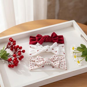 May include: Three baby headbands with bow accents. The top headband is a solid red, the middle one is white with red cherry prints, and the bottom one is white with red heart prints. They are arranged on a white tray with decorative berries and flowers.