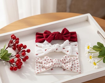Op de afbeelding: Drie baby haarbanden met strikaccenten. De bovenste haarband is effen rood, de middelste is wit met rode kersenprints en de onderste is wit met rode hartjesprints. Ze zijn gerangschikt op een witte dienblad met decoratieve bessen en bloemen.