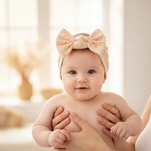 May include: A baby wearing a peach-colored headband with a large bow and small polka dots. The baby is being held by someone, with the baby's eyes open and looking at the camera. The background is soft and out of focus.