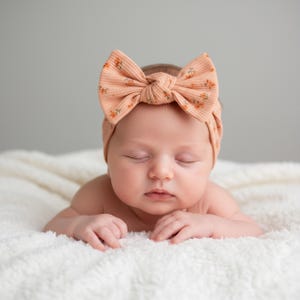 May include: A peach-colored baby headband with a large bow and a floral pattern. The headband is ribbed and worn by a baby lying on a white, fluffy blanket. The baby has their eyes closed.