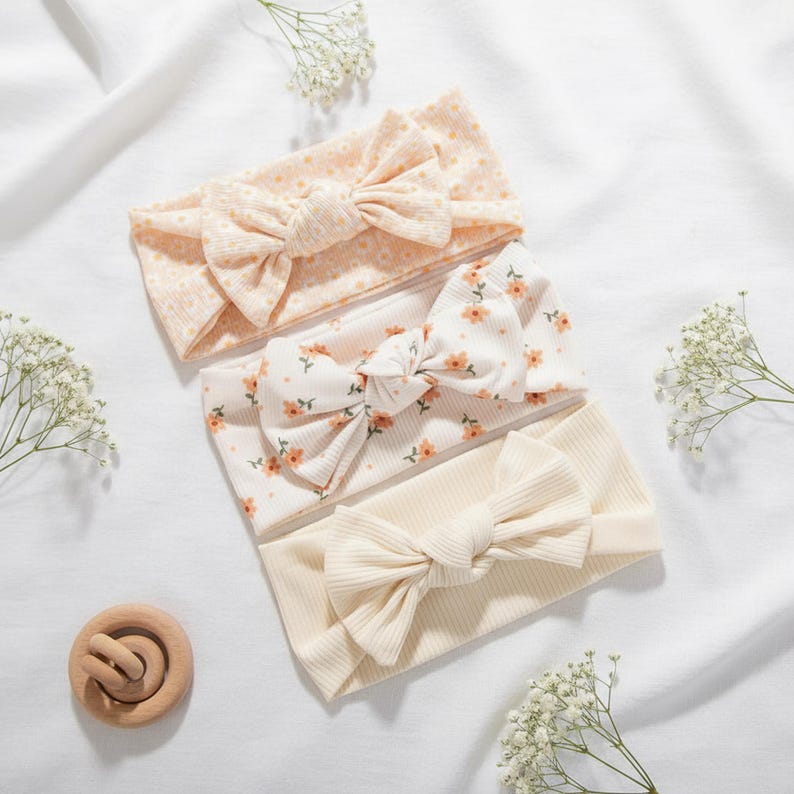 May include: Three baby headbands with large bow accents. The headbands are in shades of cream and peach, with floral and dotted patterns. A wooden teething ring is also visible.