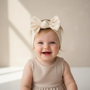 May include: A smiling baby wearing a beige ribbed headband with a large bow. The baby is also wearing a beige sleeveless dress. The background is a soft, neutral color.