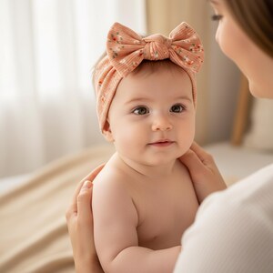May include: A baby wearing a peach-colored ribbed headband with a large bow. The bow is decorated with a small floral pattern in shades of pink and white. The baby is being held by someone out of frame.
