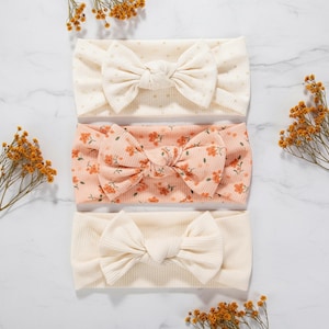 May include: Three headbands with bow details. The top headband is cream with small polka dots. The middle one is peach with a floral pattern. The bottom headband is cream with a ribbed texture. Each headband features a bow in the centre.
