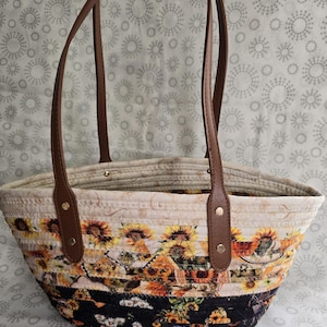 Puede incluir: Un bolso tipo tote hecho de cuerda enrollada con un estampado de girasoles en tonos amarillo, naranja y negro. El bolso tiene dos correas de hombro de piel sintética marrón sujetas con remaches metálicos dorados y un forro interior crema.