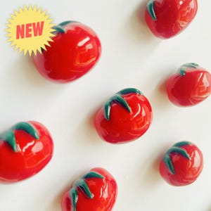 May include: A collection of glossy red tomato-shaped ornaments with dark green stems. One tomato in the upper left corner features a yellow starburst graphic with the word NEW printed in bold red capital letters across the center of the star.