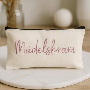 Könnte beinhalten: Eine rechteckige Kosmetiktasche aus cremefarbenem Canvas mit einem schwarzen Reißverschluss und dem Wort "Mädelskram" in rosa Glitzerschrift. Die Tasche liegt auf einer weißen Marmoroberfläche.