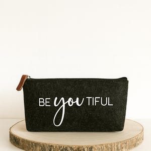 Könnte beinhalten: Ein schwarzes Filz-Reißverschlussetui mit den Worten "BE you TIFUL" in weißer Schrift. Das Etui hat einen braunen Leder-Zuglasche und wird auf einer Holzscheibe präsentiert.