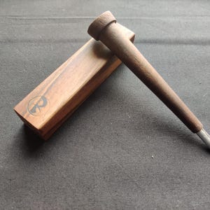 May include: A wooden tool with a cylindrical handle and a metal tip, resting on a rectangular wooden box. The box has a logo on it. The items are made of wood and metal.
