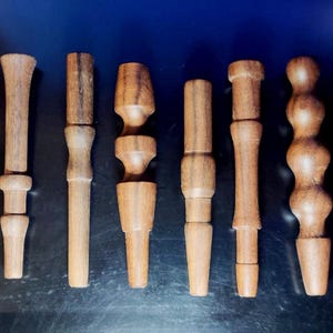 May include: A collection of various wooden bottle stoppers, each with a unique design. The stoppers are crafted from light brown wood, showcasing different shapes and decorative elements. The collection includes stoppers with bulbous, cylindrical, and segmented forms.
