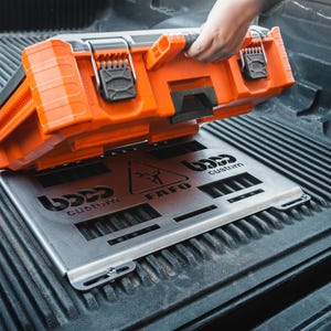 May include: An orange and black toolbox is being placed on a silver metal base with the text "BOSS custom" and "FAFO". The toolbox is resting on a black truck bed.