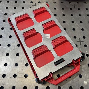 Op de afbeelding: Een rode en zilveren gereedschapsorganizer met zes rode compartimenten. De organizer heeft een rode handgreep en wielen voor gemakkelijk transport. Het metalen oppervlak heeft een rasterpatroon.