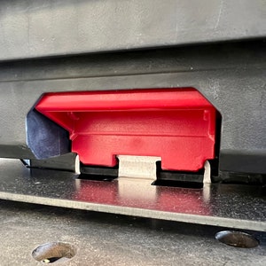 May include: A black plastic storage case featuring a prominent red latch mechanism centered on the front panel. The latch is constructed with a textured red plastic handle and a metallic silver hinge component, secured to a flat, dark gray base surface.