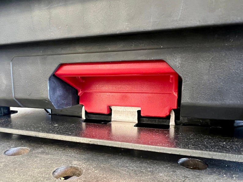 May include: A black plastic storage case featuring a prominent red latch mechanism centered on the front panel. The latch is constructed with a textured red plastic handle and a metallic silver hinge component, secured to a flat, dark gray base surface.