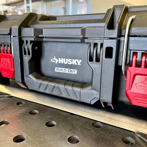 May include: A black Husky Build-Out tool box featuring a central carrying handle and two bright red locking latches on the front. The Husky logo and the text Build-Out are clearly visible on the front panel of the durable plastic storage case.