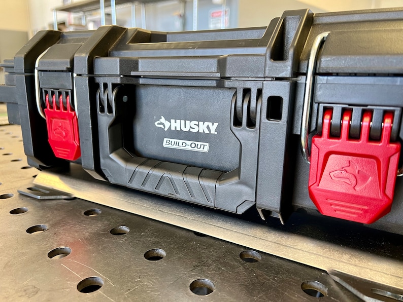 May include: A black Husky Build-Out tool box featuring a central carrying handle and two bright red locking latches on the front. The Husky logo and the text Build-Out are clearly visible on the front panel of the durable plastic storage case.