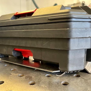 May include: A black plastic tool box with a red handle and red latch mechanism. The rectangular container features a sturdy, molded design with a flat lid and is mounted on a silver-colored metal bracket base with visible mounting slots.