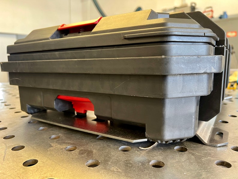 May include: A black plastic tool box with a red handle and red latch mechanism. The rectangular container features a sturdy, molded design with a flat lid and is mounted on a silver-colored metal bracket base with visible mounting slots.
