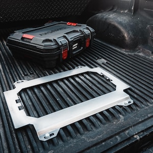 May include: A black Husky toolbox with red accents and a silver metal frame are in the bed of a truck. The frame is rectangular with bolt holes. The truck bed is black with ribbed lines.