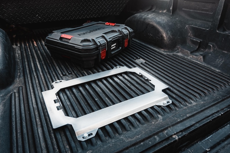 May include: A rectangular silver metal frame with four corner mounting points and integrated side brackets, positioned on a black ribbed truck bed liner next to a black and red plastic tool case with the word Husky printed on the lid.