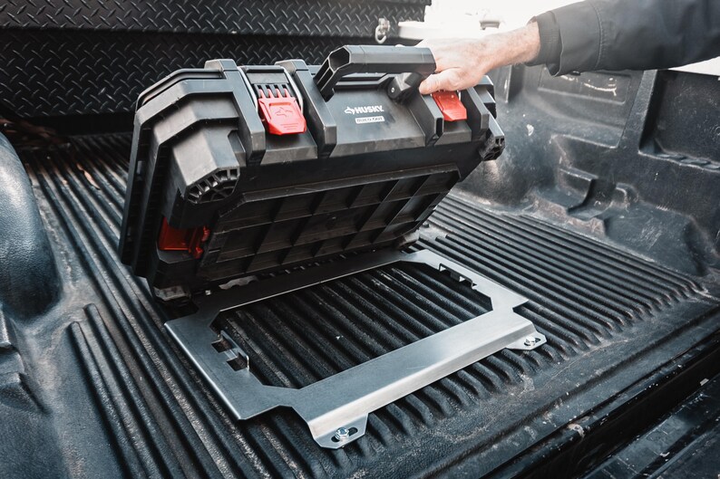 May include: A black Husky brand plastic tool box with red locking latches and a top handle, positioned above a silver metal mounting bracket. The tool box is being lifted to reveal the rectangular metal frame secured to a ribbed truck bed surface.