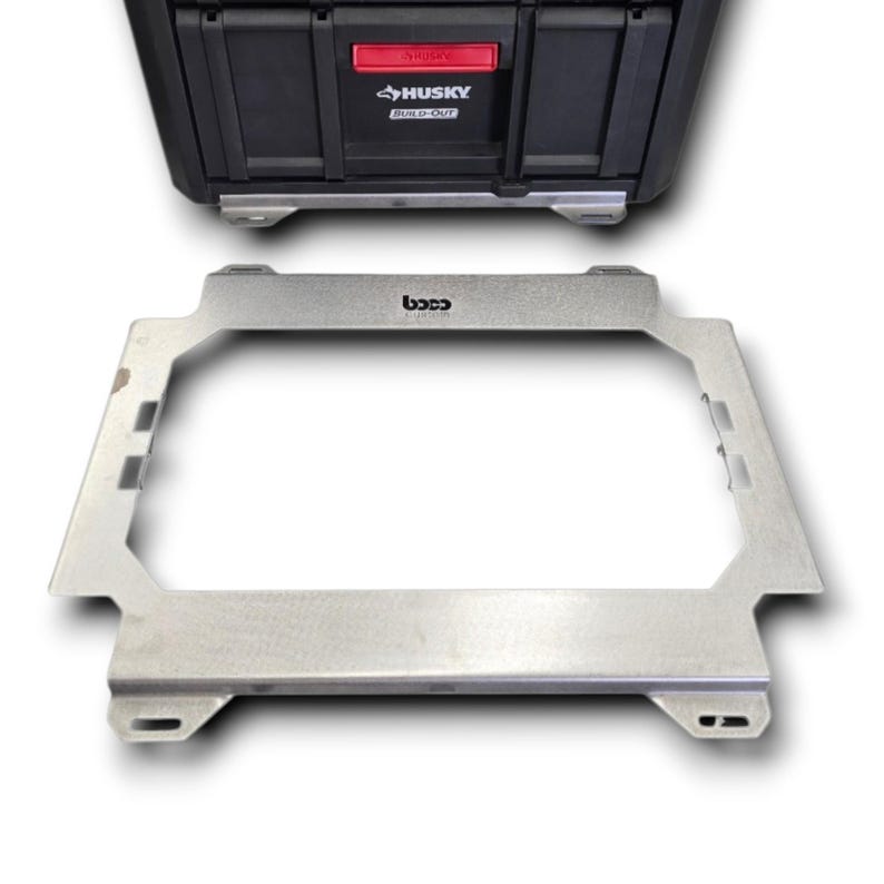 May include: A rectangular silver metal mounting bracket with a central cutout and integrated side slots, featuring the text "boco" printed in black on the top edge, positioned in front of a black Husky Build-Out storage case with a red latch.