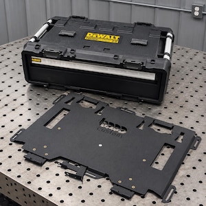 Puede incluir: Una caja de herramientas negra DeWalt ToughSystem 2.0 con un asa plateada y una placa de montaje de metal negro separada que presenta el texto cortado con láser "bass custom" en el centro, sobre una superficie de metal perforada con un enchufe.