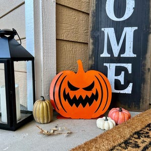May include: A black lantern with a white candle sits next to a wooden sign that says "HOME". An orange pumpkin with a carved face and several decorative pumpkins in various colors are displayed on a porch, creating a festive Halloween scene.