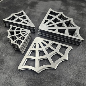 May include: A collection of silver-colored metal corner accents shaped like spider webs. The set includes multiple stacks of these triangular, cut-out web designs in varying sizes, featuring a flat, metallic finish and geometric patterns.