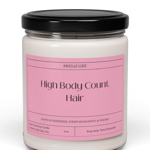 May include: A white soy candle in a glass jar with a black lid. The label is pink and says "Smells Like High Body Count Hair" with the text "Hints of frizziness, stripy highlights, & volume". The candle is 9 oz and hand-poured in Salt Lake City.