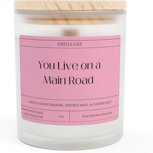Puede incluir: Un tarro de vela blanco con una tapa de madera. La etiqueta dice "Smells Like You Live on a Main Road" con el texto "Hints of chain smoking, divorce rage, & clapping back". La vela es de 11 oz.