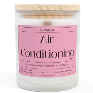 Puede incluir: Vela perfumada en un tarro de vidrio esmerilado con tapa de madera. La etiqueta rosa dice "Air Conditioning" con notas de fragancia. La vela pesa 312 gramos.