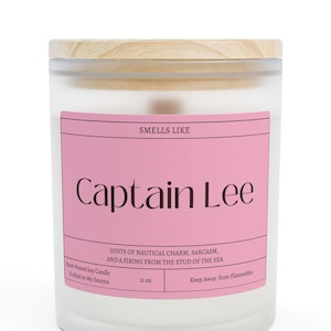 May include: A frosted glass candle with a wooden lid. The candle has a pink label that reads "Smells Like Captain Lee." The label also includes the text "Hints of nautical charm, sarcasm, and a firing from the stud of the sea." The candle is 11 oz.