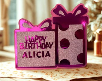 May include: A decorative birthday card shaped like two gift boxes with bows. The left box is square with glittery pink texture and the text Happy Birthday ALICIA. The right box is tall with metallic magenta edges and glittery pink polka dots.