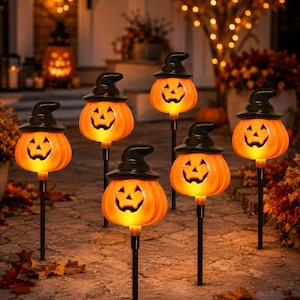 May include: Six illuminated orange pumpkin garden stakes, each featuring a black witch hat and a carved jack-o'-lantern face. These outdoor lights are mounted on thin black poles, casting a warm glow across the stone pathway at night.