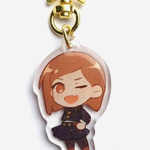 May include: A clear acrylic keychain with a gold star-shaped clasp. The keychain features a cartoon illustration of a girl with brown hair, wearing a black and gold uniform. The girl is winking and smiling.