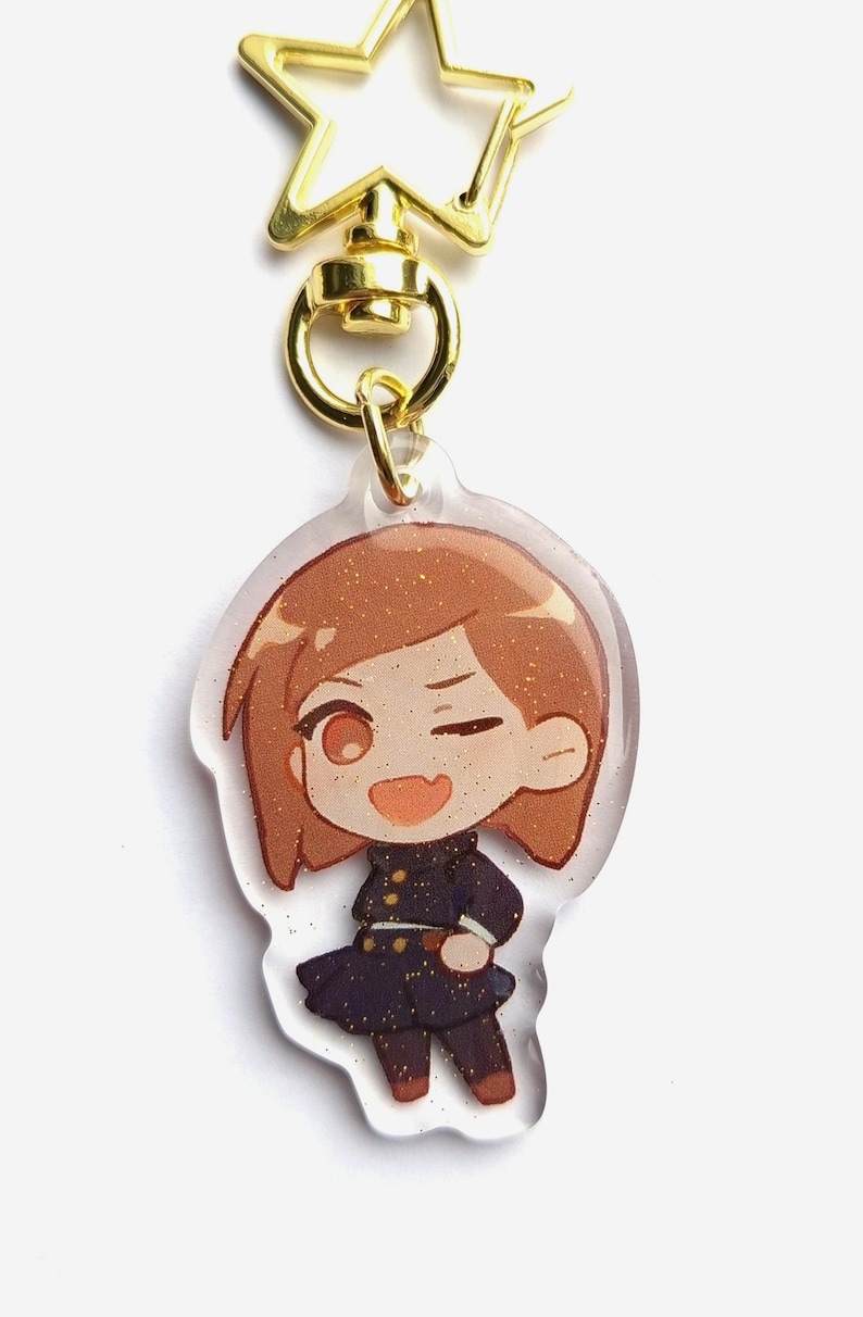 May include: A clear acrylic keychain with a gold star-shaped clasp. The keychain features a cartoon illustration of a girl with brown hair, wearing a black and gold uniform. The girl is winking and smiling.