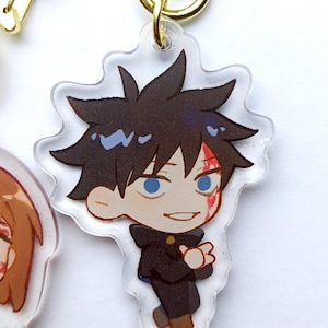 May include: A clear acrylic keychain featuring a cartoon character with black hair, blue eyes, and a red mark on their face. The character is wearing a black uniform with a white collar. The keychain is attached to a gold metal ring.