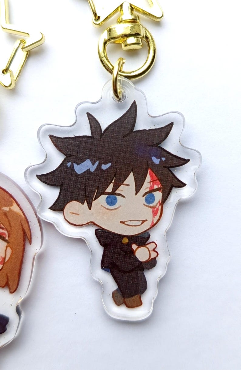 May include: A clear acrylic keychain featuring a cartoon character with black hair, blue eyes, and a red mark on their face. The character is wearing a black uniform with a white collar. The keychain is attached to a gold metal ring.