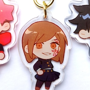 May include: A clear acrylic keychain featuring a cartoon character with brown hair, a black dress, and orange eyes. The character has a red and white design on their cheek.