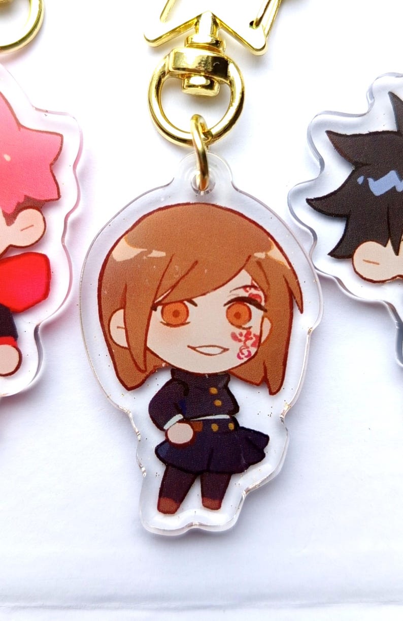 May include: A clear acrylic keychain featuring a cartoon character with brown hair, a black dress, and orange eyes. The character has a red and white design on their cheek.