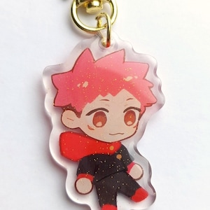 May include: A clear acrylic keychain featuring a cartoon character with red hair, a red scarf, and a black and gold uniform. The keychain is attached to a gold star-shaped clasp.