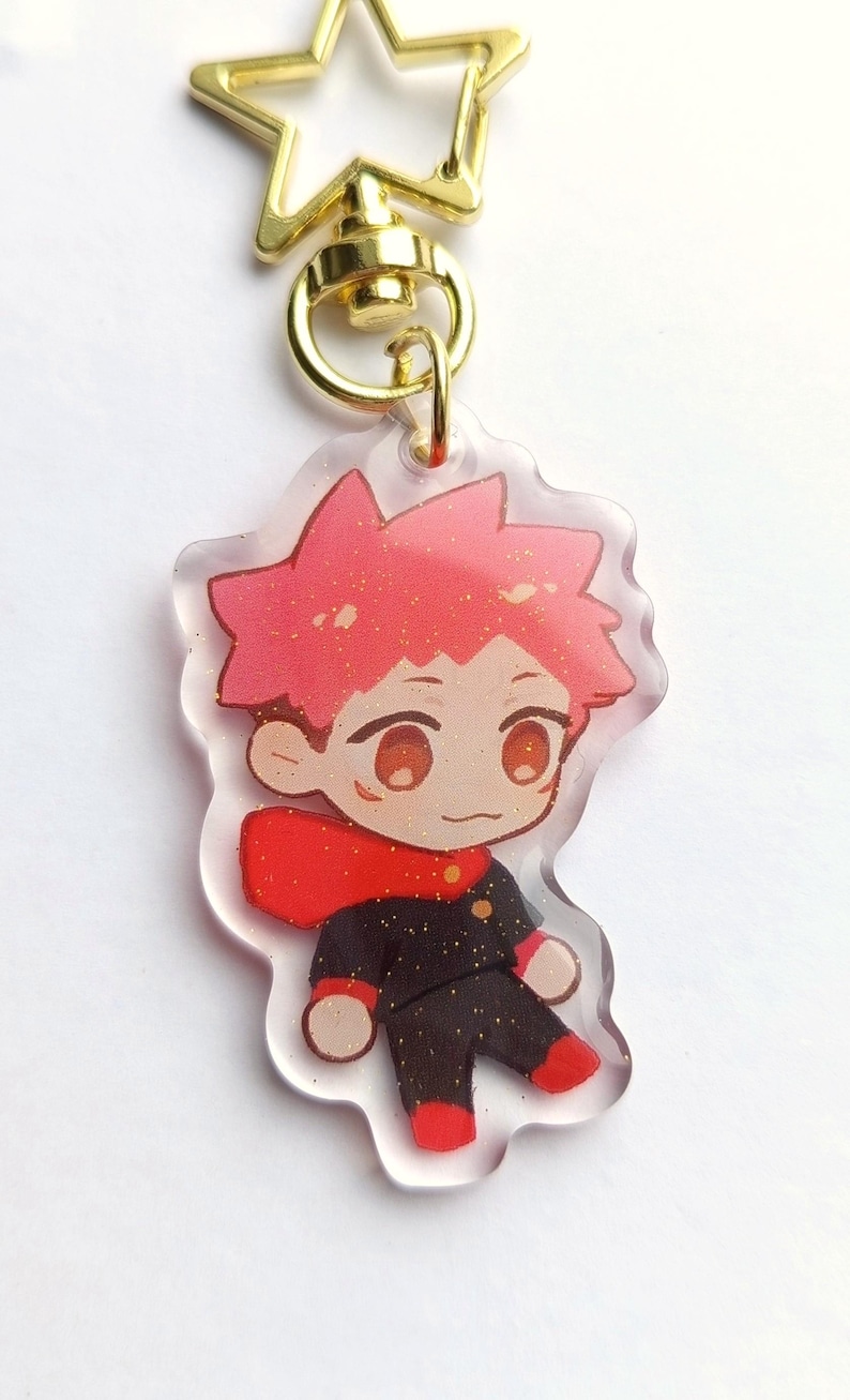 May include: A clear acrylic keychain featuring a cartoon character with red hair, a red scarf, and a black and gold uniform. The keychain is attached to a gold star-shaped clasp.