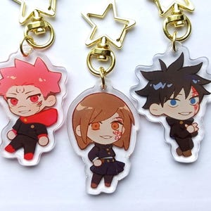 May include: Three anime character keychains with gold star-shaped charms. The characters are a girl with brown hair and a red and black outfit, a boy with black hair and a black and white outfit, and a girl with pink hair and a red and black outfit.