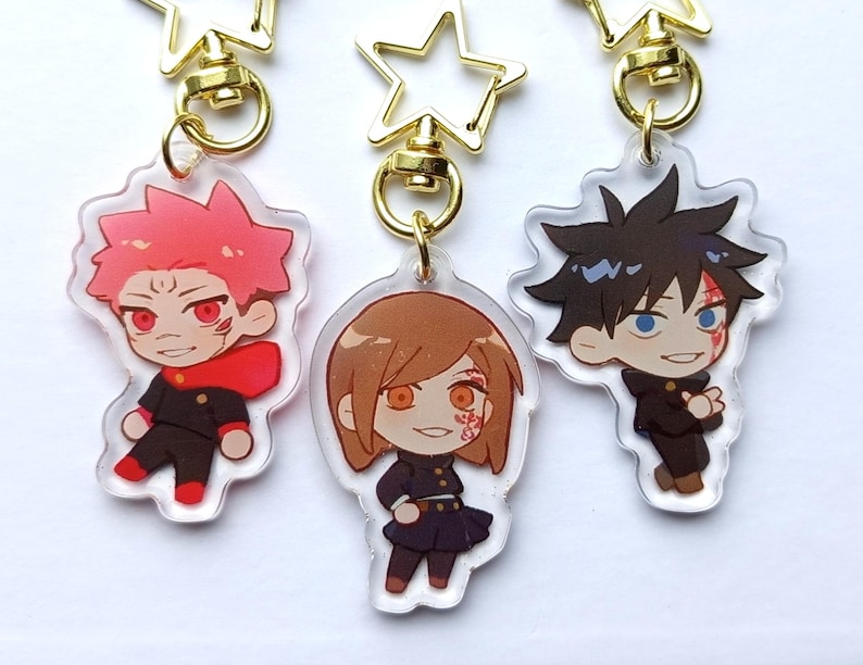 May include: Three anime character keychains with gold star-shaped charms. The characters are a girl with brown hair and a red and black outfit, a boy with black hair and a black and white outfit, and a girl with pink hair and a red and black outfit.
